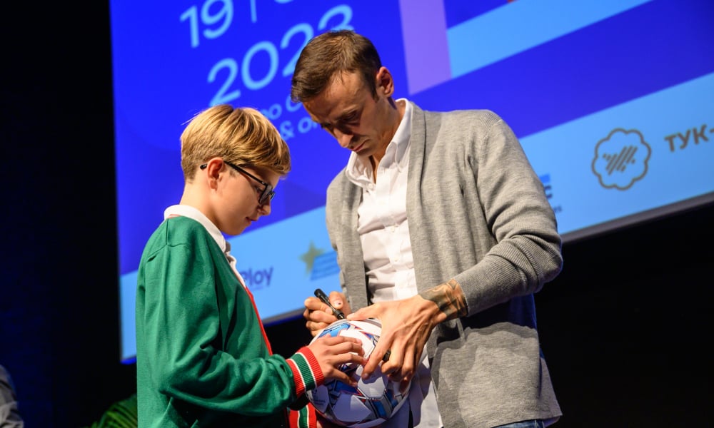 Dimitar Berbatov signs a soccer ball held by a boy in glasses and green jacket.