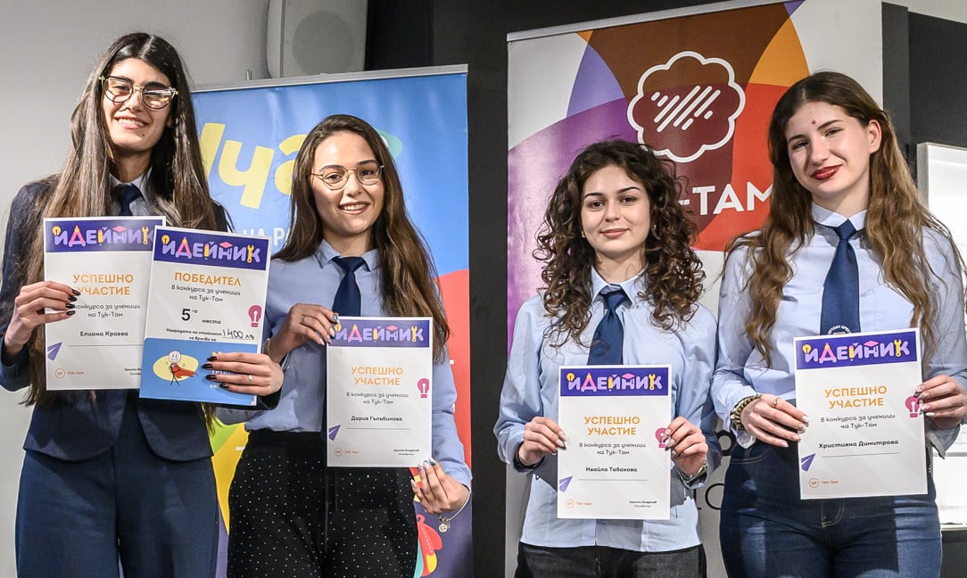 Four young women holding certificates and smiling at an award event with colorful banners behind them.