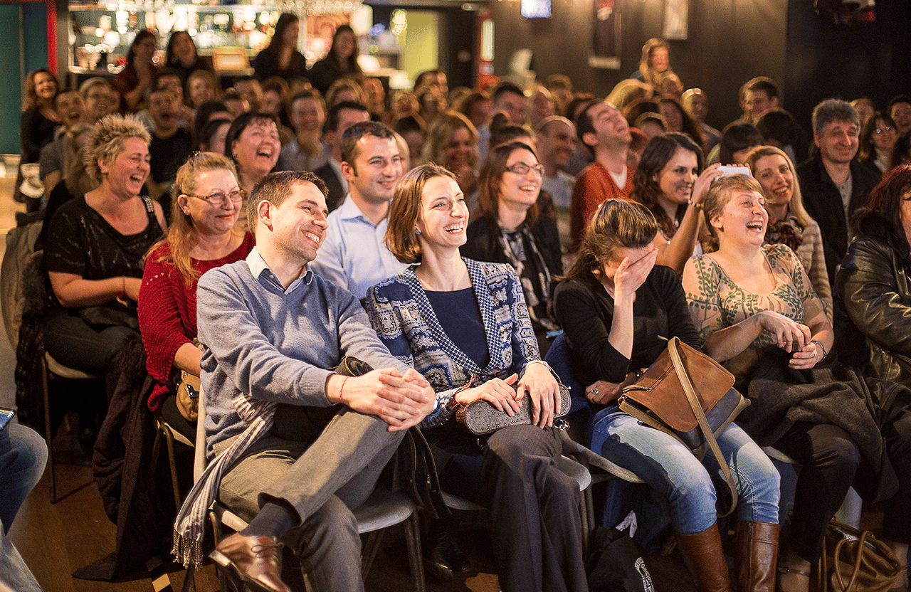 Audience sitting closely together laughing and enjoying an comedy event indoors.