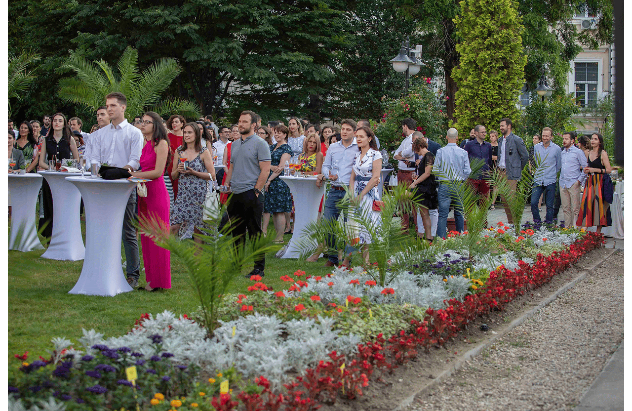 Group of people attending an outdoor garden event, standing and socializing near tall white cocktail tables.