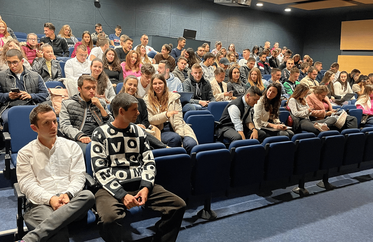 Audience of mixed young adults seated in a lecture hall with blue chairs and dark walls.
