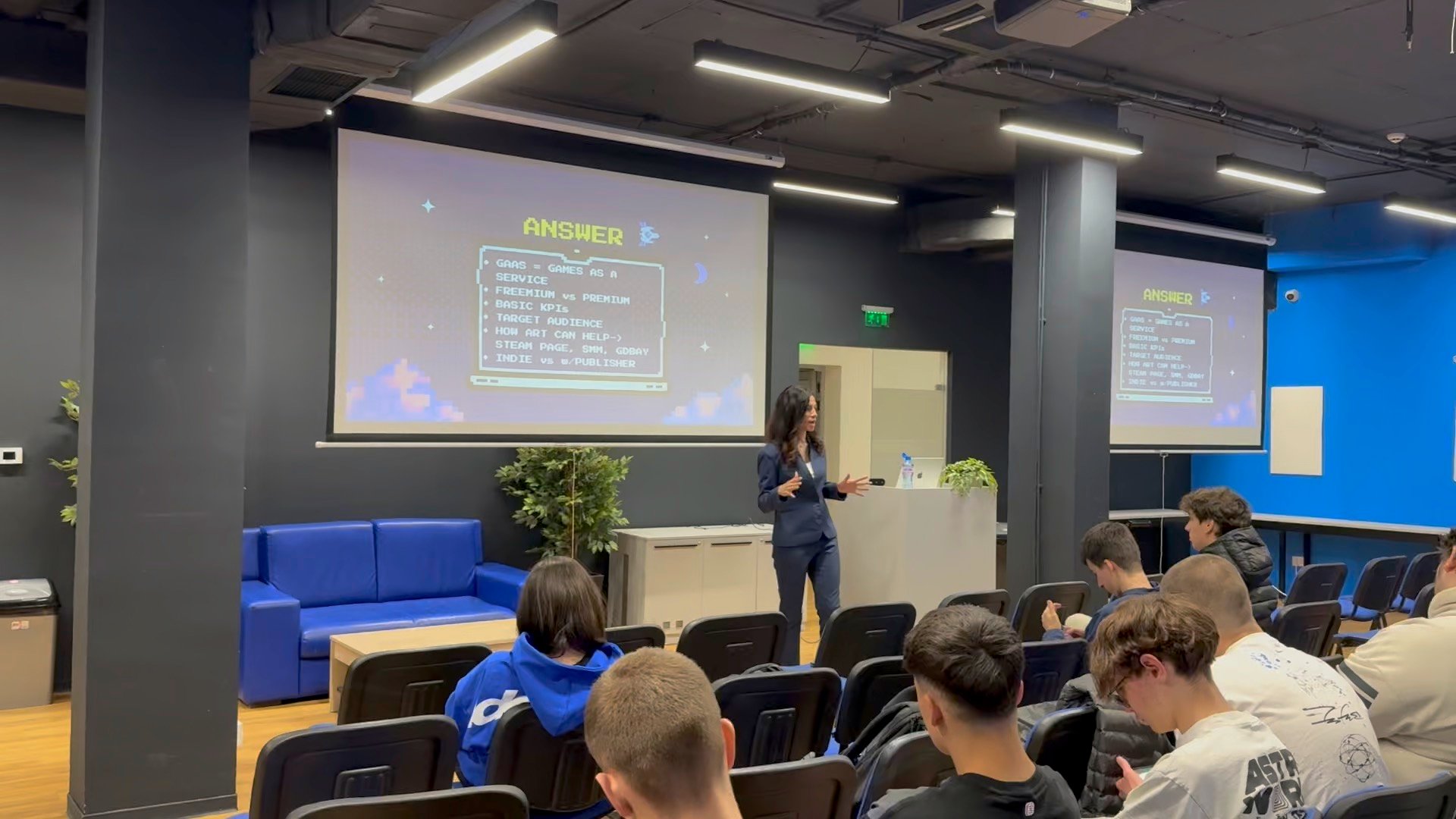 Woman in blue suit speaking to seated audience in classroom with slides about games.