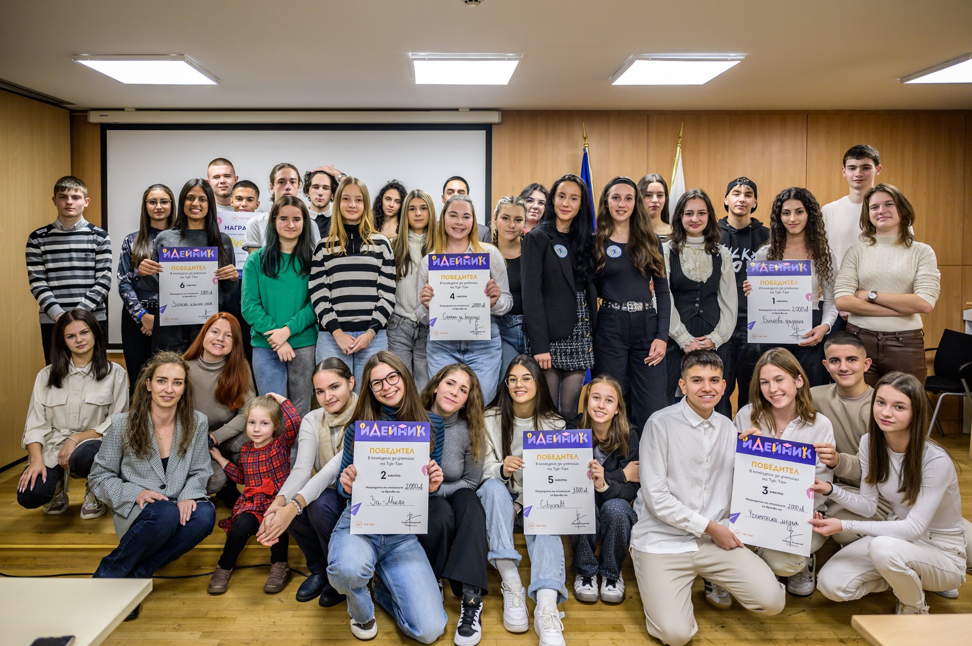 Group of young award winners and mentors posing indoors, some holding certificates