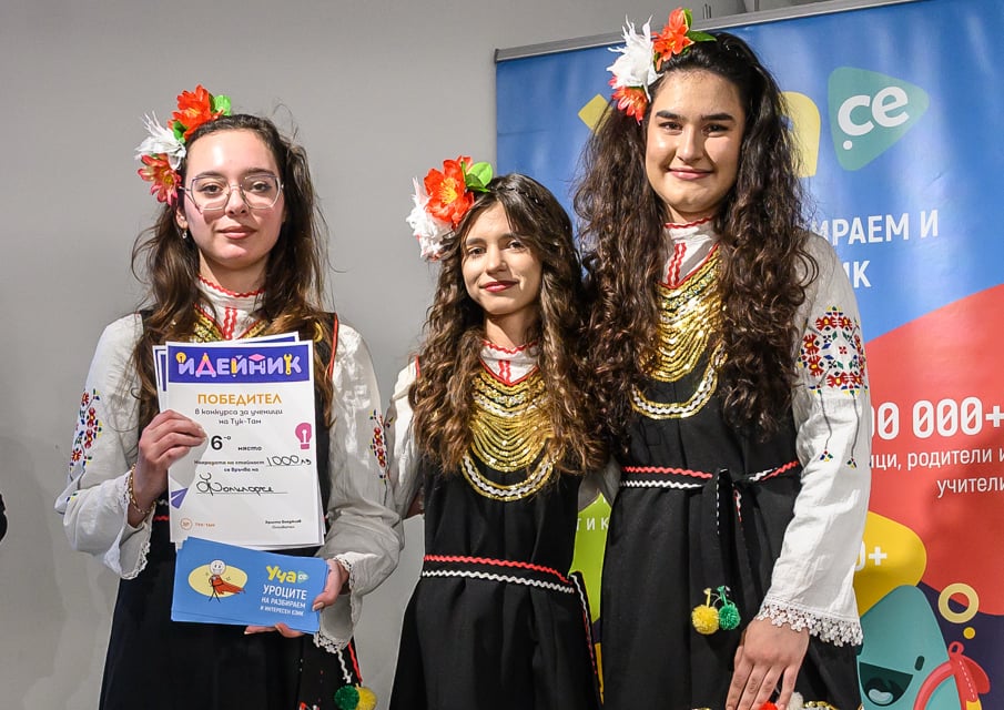 Three women in traditional embroidered dresses with floral headpieces; one holds a certificate and envelope.