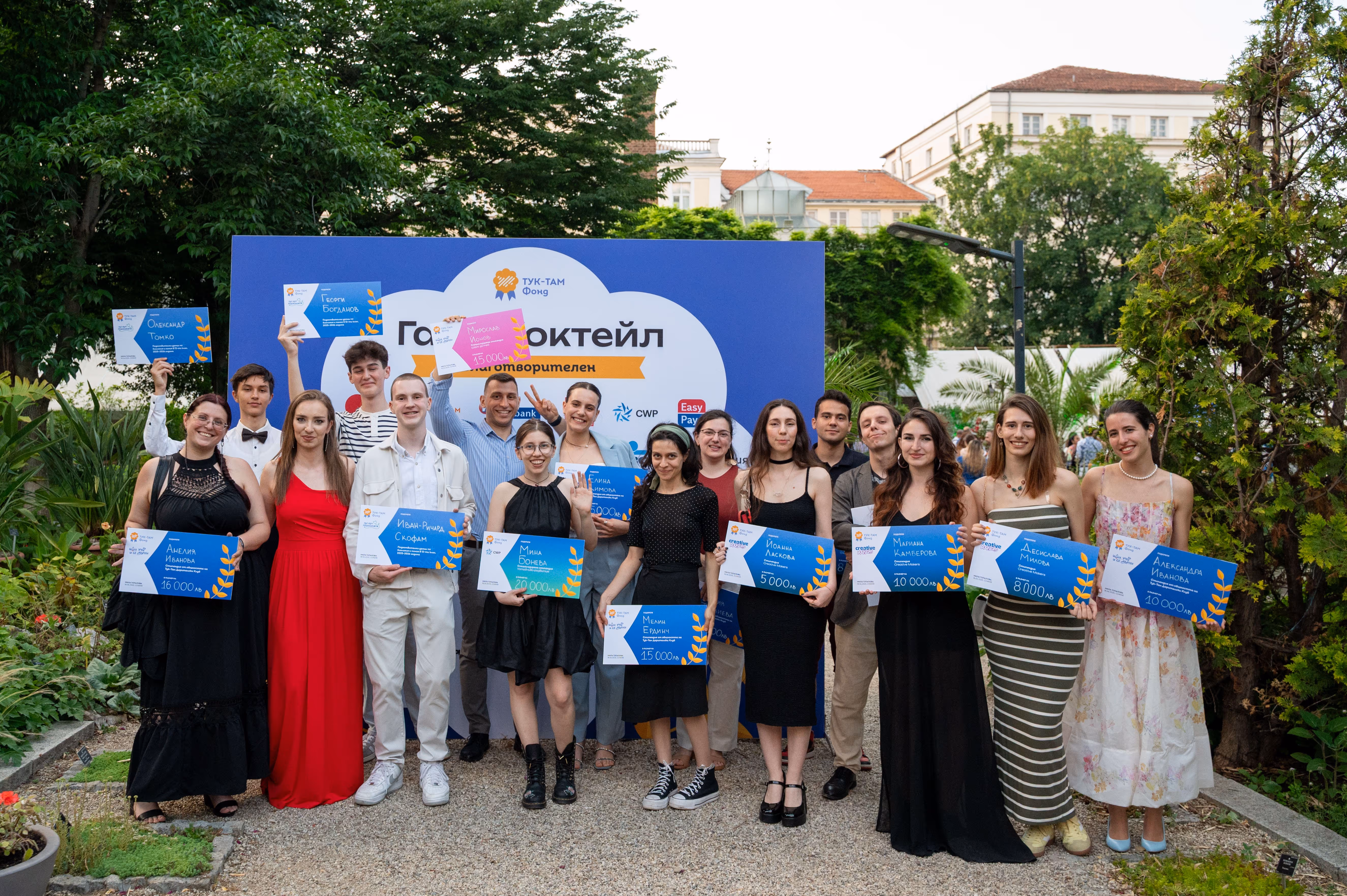 Group of young adults holding award certificates posing outdoors in front of an event banner.